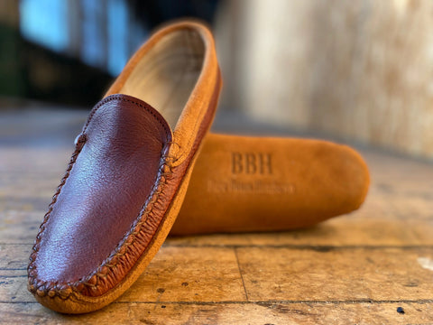 A close-up of BBH’s Northeaster’ moccasin slippers in brown and tan premium leather, one upright and one flat on a wooden floor, features the BBH emboss—celebrating fine American craftsmanship.