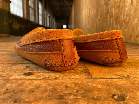 A low-angle close-up of BBH Northeaster’ handsewn moccasin slippers, crafted from premium leather, displayed on a worn wooden floor in a sunlit rustic hallway with large windows and textured walls.