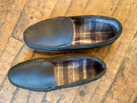 A pair of BBH Northeaster’ black handsewn moccasin slippers, made from premium leather with plaid fleece interiors, rest side by side on a wooden floor—highlighting authentic American craftsmanship.