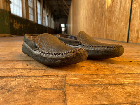 A pair of BBH Northeaster’ handsewn moccasin slippers made from premium leathers rests on a worn wooden floor in a hallway, natural light streaming through tall windows on the left.