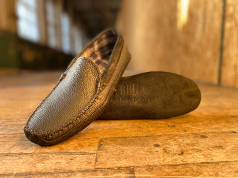 A pair of BBH Northeaster’ handsewn brown leather moccasin slippers is displayed on a wooden floor—one upright, one flat—set against a blurred indoor background that highlights American craftsmanship.