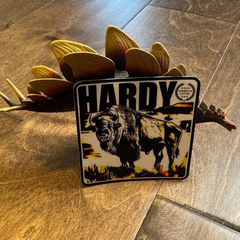 The American Lictor Sticker: Bison - Hardy (3 x 3) stands on a wooden floor, with a brown artificial flower behind it. Sunlight highlights this bold sticker, casting dynamic shadows and emphasizing its American icon design.