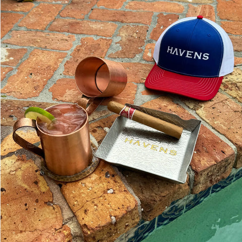 A copper mug with a lime wedge, a cigar on a stainless steel Havens Luxury Metals ash tray, another copper mug on its side, and a red, white, and blue HAVENS cap rest on the brick edge beside the pool.