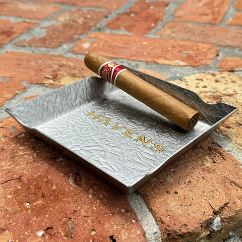 A brown cigar rests on the Ash Tray - Stainless Steel by Havens Luxury Metals, featuring gold HAVENS engraving, set on a rustic brick surface.