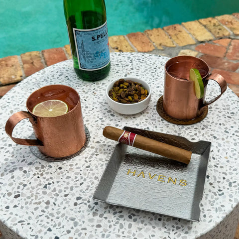 A round table by a pool holds two copper mugs with lime slices, a green San Pellegrino bottle, a bowl of pistachios, and a cigar resting on the Ash Tray - Stainless Steel from Havens Luxury Metals.