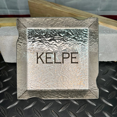 A Havens Stainless Steel Ashtray by Havens | Luxury Metals, handcrafted in the USA, with a textured silver square design, rests on a diamond plate surface with a white foam block in the background.