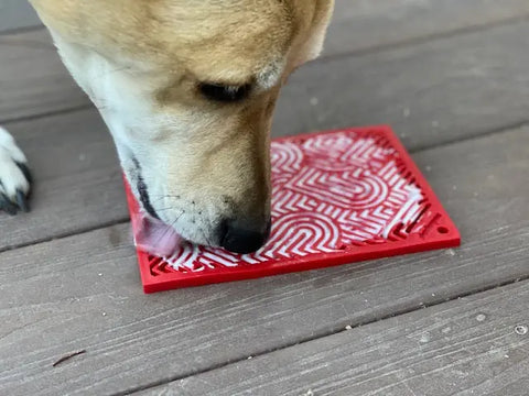 A dog enjoys a treat on the SodaPup Heart Design Love EMat Lick Mat, a red and white enrichment toy, while lying on a wooden floor.