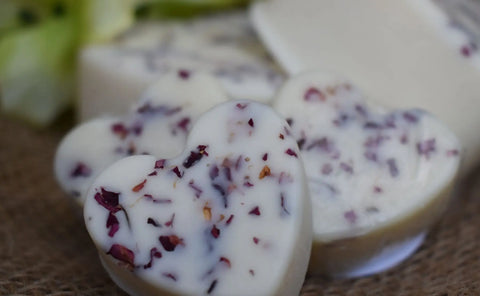 Close-up of Bathsheba Body Works artisan-crafted Moisture Bar - Love Me, heart-shaped with dried flower petals—ideal for mature skin—displayed on burlap with blurred soap bars and greenery in the background.