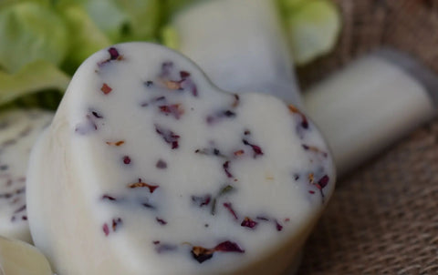 A close-up of Bathsheba Body Works Moisture Bar – Love Me, an artisan-crafted, heart-shaped bar with colorful dried flower petals, placed on a woven surface with green leaves. Ideal for nourishing mature skin.