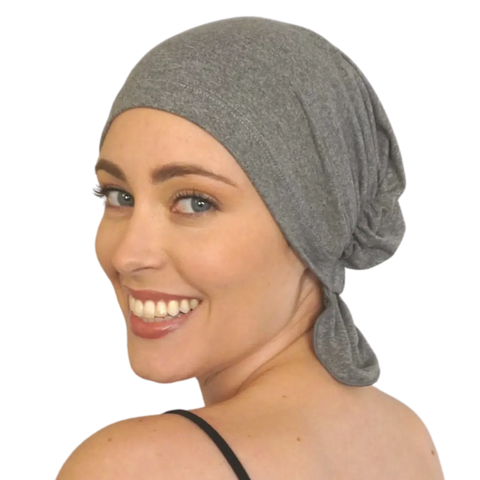 A smiling woman with blue eyes wears the Chemo Beanies® Patricia grey chemo scarf and a black strap top, looking over her shoulder against a plain background.