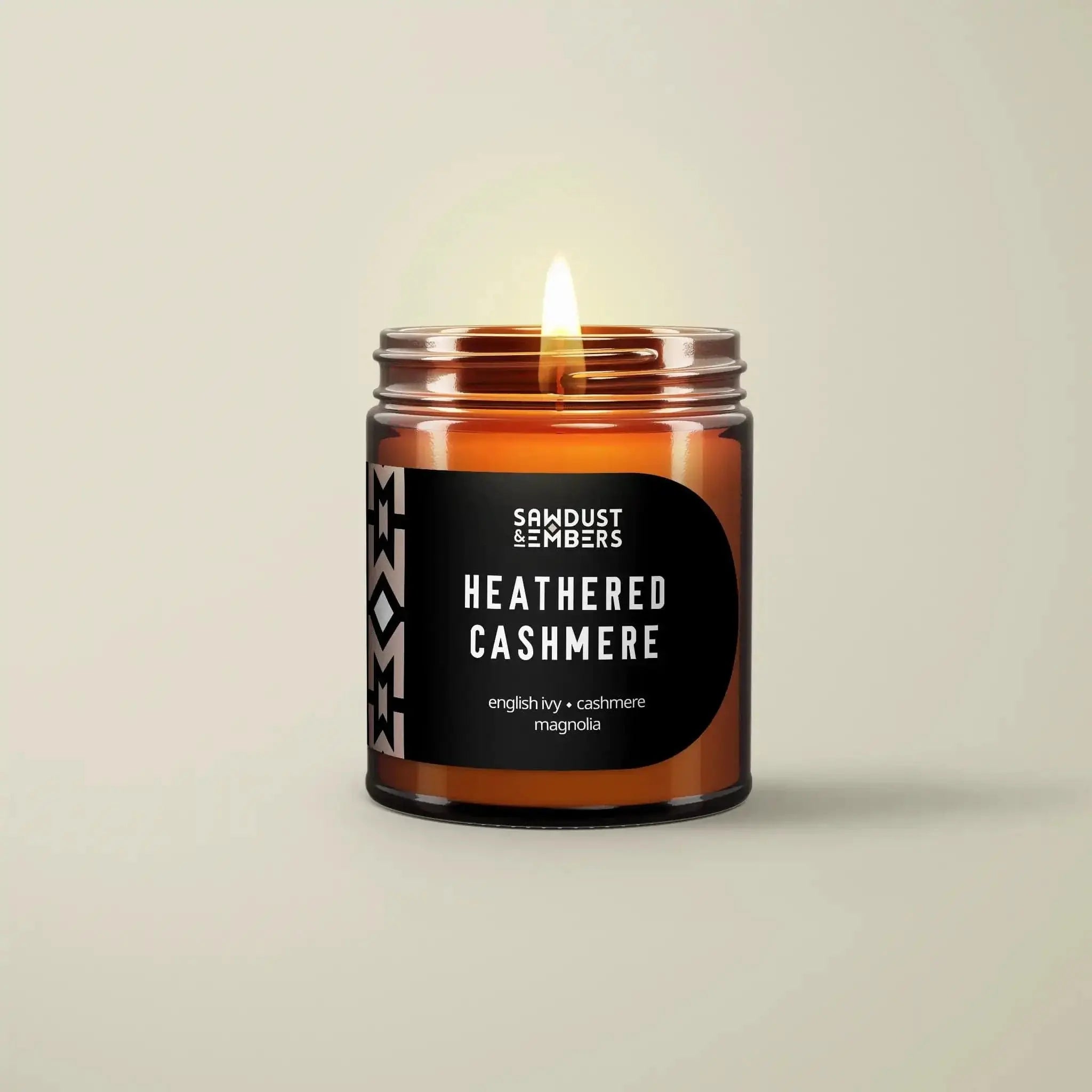 A brown glass jar Heathered Cashmere - Signature Candle by Sawdust & Embers, lit with a warm flame. The label lists English ivy and cashmere magnolia scents—ideal for adding a touch of luxury to your home. Light beige background.