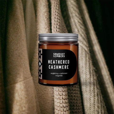 A brown Heathered Cashmere - Signature Candle by Sawdust & Embers sits before soft beige and brown sweaters, creating a cozy home fragrance scene.