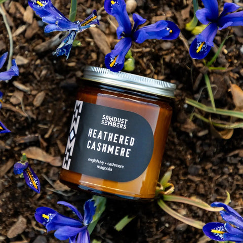 A brown glass Heathered Cashmere - Signature Candle by Sawdust & Embers, set on soil amid blooming purple and yellow flowers, features a silver lid and label with home fragrance notes of English ivy, cashmere, and magnolia.