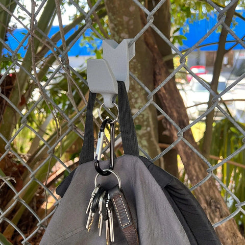 A gray bag with keys hangs from the Chatelet Manufacturing Carabiner Fence Holder, which is attached to a white plastic hook on a chain-link fence outdoors. Trees and parked cars are visible in the background.