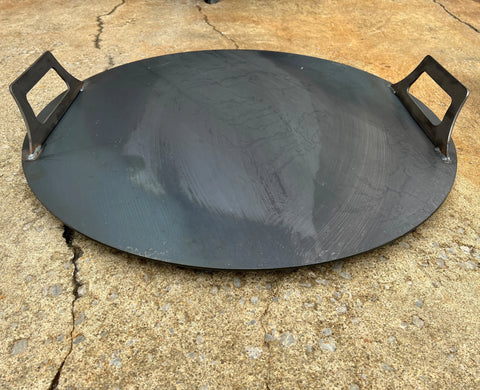 An Ohio Flame Flat Fire Pit Lid (Made in USA) with two rectangular handles rests on a rough, cracked concrete surface.
