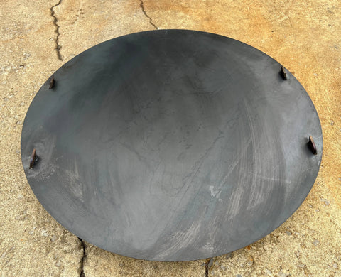 An Ohio Flame Flat Fire Pit Lid (Made in USA), large, black, and round with four small metal tabs around the edge, rests on a rough concrete surface.