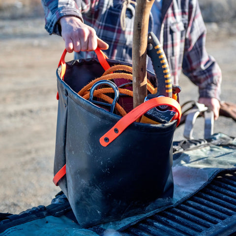 A person stands by a truck bed outdoors, holding the eqpd DirtBag—a rugged tote with orange handles—packed with tools and ropes. Dressed in a plaid shirt, theyre ready for trail building.