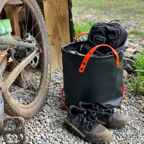 A pair of muddy cycling shoes rest on gravel next to the eqpd DirtBag, a rugged tote with orange handles filled with clothes, beside a dirty mountain bike leaning against wood. Grass and rocks complete the background scene.
