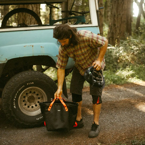 A long-haired, bearded man stands by a blue truck in the woods, holding a black helmet and reaching for an eqpd DirtBag with orange handles. A bicycle sits in the truck bed—a classic scene for trail builders.