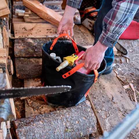 Wearing a plaid shirt, someone lifts the eqpd DirtBag tote packed with tools and a fuel can, surrounded by cut firewood and logs on a wooden surface—ideal gear for trail builders.