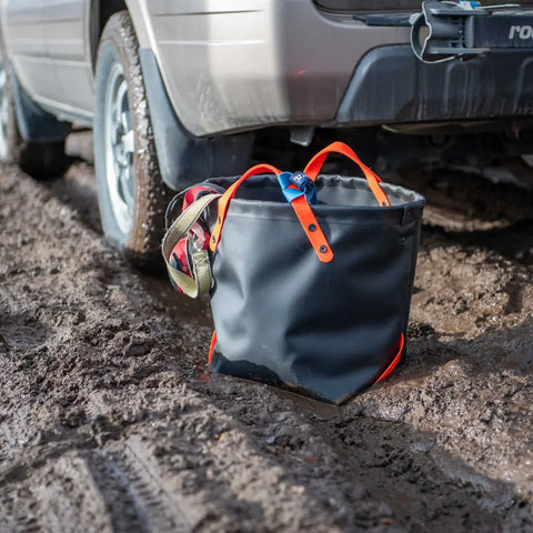 The eqpd DirtBag is a rugged, black, waterproof 6+ gallon tote with orange handles, seen on muddy ground behind a parked car. A plaid cloth peeks out, and tire tracks in the mud highlight its utility for trail builders.