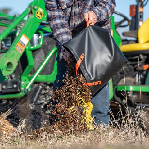 Wearing jeans and a plaid shirt, a person dumps an eqpd DirtBag of mulch onto the ground near a green tractor—ideal for trail builders and outdoor work.