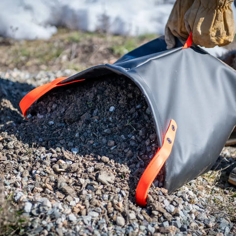 Wearing gloves, a person pours soil from the eqpd DirtBag—a rugged tote with bright orange handles—onto the ground outdoors, making it ideal for trail builders.