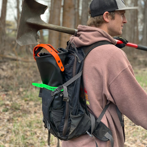 Outdoors in a forested area, a person in a brown hoodie and cap carries a shovel and wears an eqpd DirtBag tote with tools strapped to their backpack—a scene typical for dedicated trail builders.