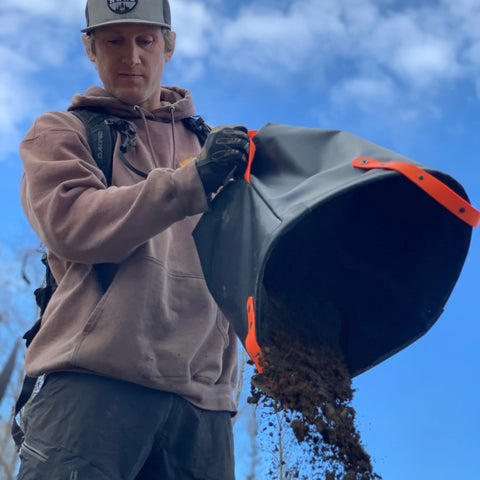 Outdoors under a blue sky, a person in a hoodie, gloves, and cap pours soil from the rugged 6+ gallon eqpd DirtBag—a must-have for trail builders.
