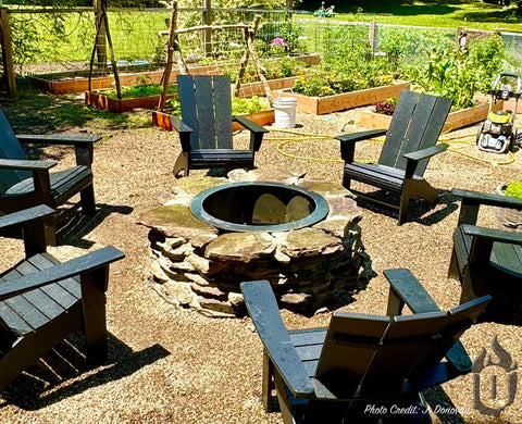 Six black wooden chairs circle a sunny backyard garden’s centerpiece: the Ohio Flame Heavy Gauge Fire Ring - Round (Made in USA), surrounded by raised beds and lush greenery.