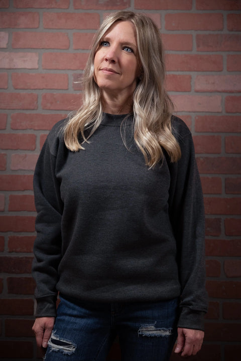 A woman with long blonde hair, dressed in the Proudly USA Crewneck Sweatshirt (Heather Charcoal) and jeans, stands before a brick wall, gazing slightly upward with a thoughtful expression.
