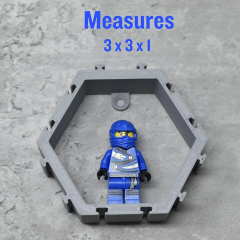 A blue LEGO ninja minifigure is displayed in a Chatelet Manufacturing Hexagon Wall Display Compatible With Lego Figures on a gray surface. Blue text above reads Measures 3 x 3 x 1.