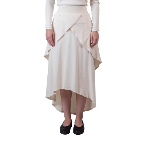Someone models the Sylva Skirt by Spiritex | Sustainable Apparel—a cream, eco-friendly skirt with layered panels and a fitted waistband—paired with a long-sleeve top and black shoes against a white background.