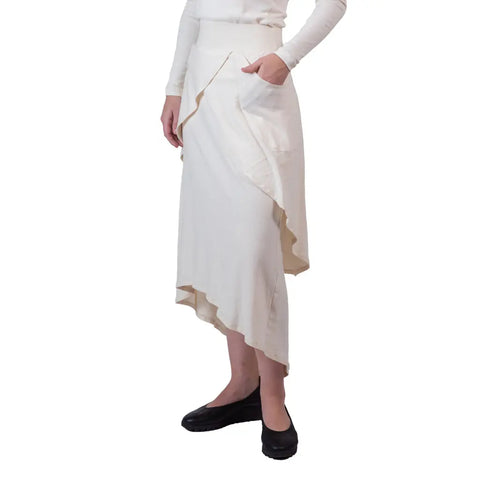 A person wears a long-sleeved white top and the Sylva Skirt by Spiritex | Sustainable Apparel, featuring a ruffled edge and pocket, paired with black flats, standing against a plain white background.