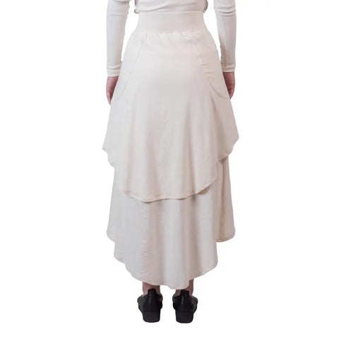 Back view of a person wearing the Sylva Skirt by Spiritex | Sustainable Apparel—an eco-friendly, cream A-line skirt with layered panels and an elastic waistband—paired with black shoes, standing against a white background.