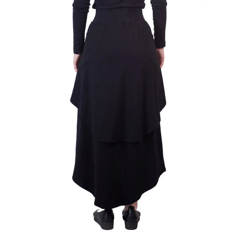 A person wearing the Sylva Skirt by Spiritex | Sustainable Apparel—an eco-friendly, long, layered black skirt—and black shoes stands with their back to the camera against a white background.