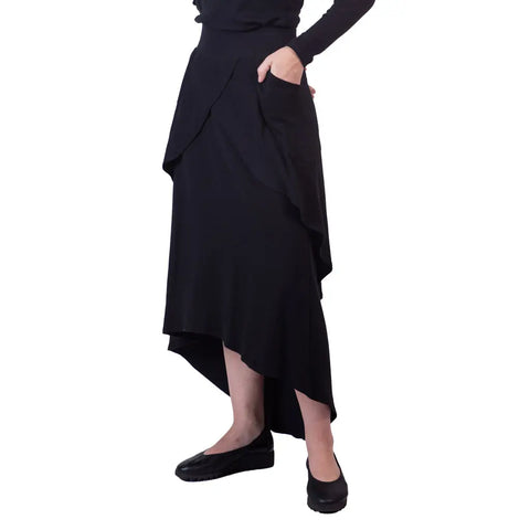 A person wears the Sylva Skirt by Spiritex | Sustainable Apparel—an organic black cotton A-line midi skirt with pockets, asymmetrical hem (shorter front, longer back)—paired with a black long-sleeve top and shoes against a white background.