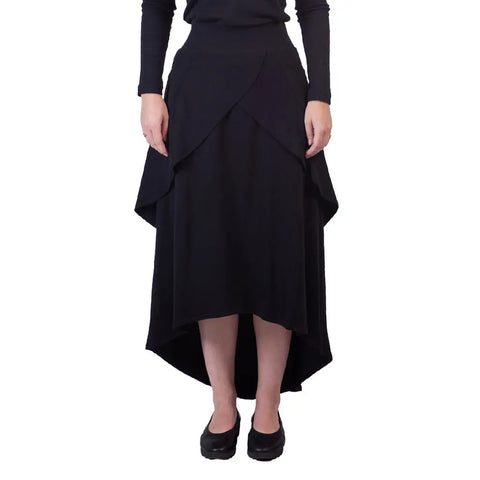 Wearing the Sylva Skirt by Spiritex | Sustainable Apparel, this eco-friendly black A-line skirt features layered panels and a high-low hemline, paired with a black top and shoes against a white background.