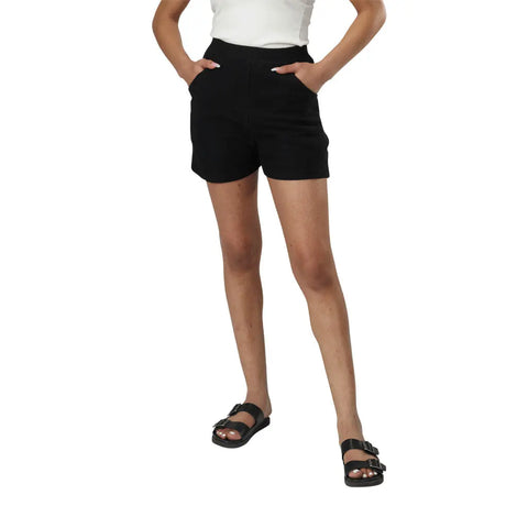 A person models the Spiritex | Sustainable Apparel Betty Hi Waist Shorts in black, paired with a white top and black sandals, standing with hands in pockets against a plain white background.