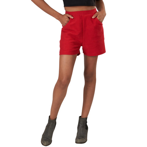 A person wearing a black crop top, Spiritex | Sustainable Apparel Betty Hi Waist Shorts in vintage-inspired bright red with pockets, and gray ankle boots stands with hands in pockets against a white background.