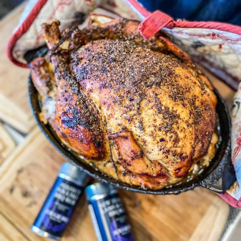 A golden brown roasted chicken sits in a skillet beside two jars from Gustus Vitae’s Complete Holiday Mains & Sides Meal Kit, featuring 10 all-natural gourmet seasonings—perfect for every occasion. An oven mitt is partially visible in the background.