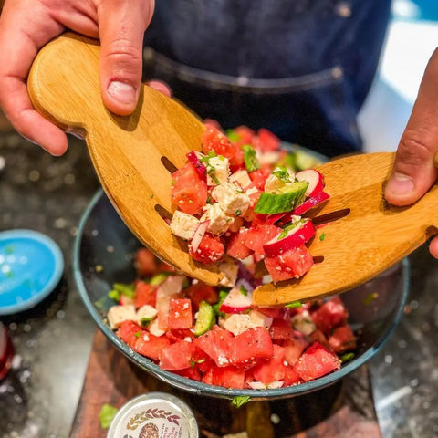 Using wooden salad servers, a person mixes watermelon salad with feta, cucumber, herbs, and Gustus Vitae Complete Holiday Mains & Sides Meal Kit’s all natural gourmet seasonings in a bowl on the kitchen counter.
