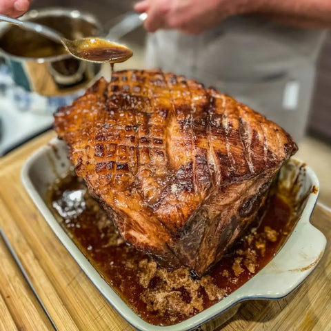 A juicy, roasted pork with crispy skin sits in a dish as a spoon drizzles artisanal sauce, all enhanced by Gustus Vitaes Complete Holiday Mains & Sides Meal Kit—10 all natural gourmet seasonings perfect for festive cooking.