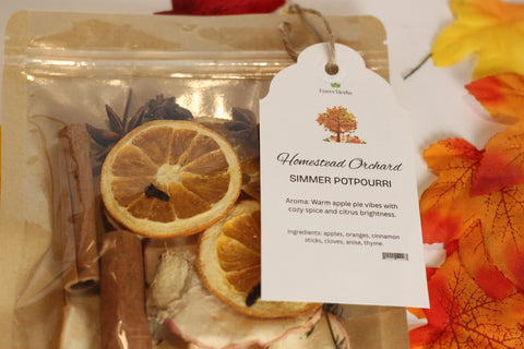 A package of Forev'Herbs Homestead Orchard Simmer Potpourri with dried orange slices, cinnamon sticks, and spices is displayed. A white tag lists aroma and ingredients. Autumn leaves in the background suggest a cozy seasonal home fragrance.