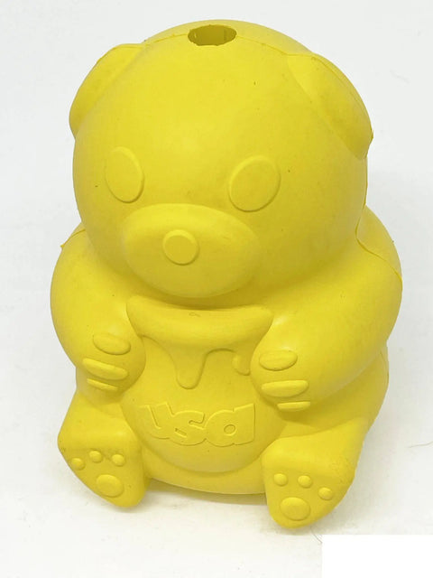 The SodaPup Honey Bear EDispenser is a yellow, bear-shaped durable rubber treat dispenser with minimal facial features, usa embossed on its belly, and a hole on top for treats. It’s pictured against a plain white background.