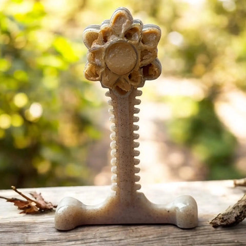 The SodaPup Honey Bone Dental Flower Tower EChew Ultra Durable Nylon Dog Chew Toy - XL stands upright on a wooden surface outdoors, shaped like a beige sunflower with a stem and base, against blurred greenery and fallen leaves.
