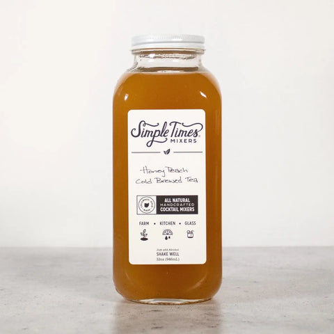 A clear glass bottle of Simple Times Mixers Honey Peach Cold Brewed Tea, made with organic black tea, filled with brownish-orange liquid sits on a gray surface against a plain white backdrop.