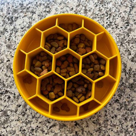 A yellow SodaPup Honeycomb Design EBowl Enrichment Slow Feeder Bowl for Dogs rests on a speckled granite countertop, with its hexagonal compartments filled with kibble to promote slower eating.