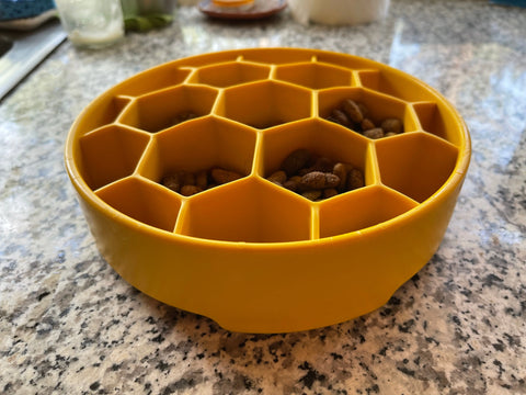 A yellow SodaPup Honeycomb Design EBowl Enrichment Slow Feeder Bowl for Dogs sits on a granite countertop, with several honeycomb sections filled with dry pet food to promote slower eating.