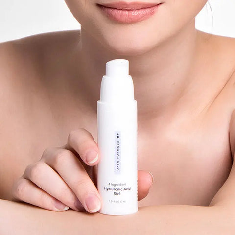 A woman with fair skin holds a white Open Formula Hyaluronic Acid Gel bottle near her face, emphasizing its dry skin benefits. Only her lower face and shoulders appear against a neutral background.